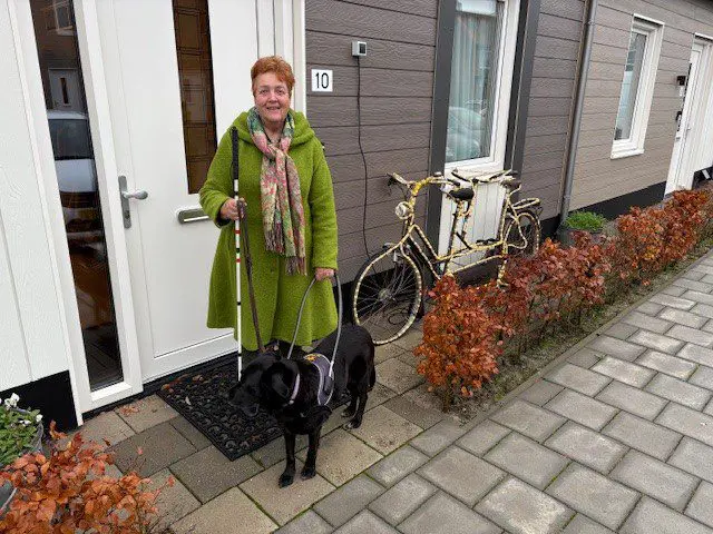Frieda en haar blindengeleidehond Reena staan voor de huisdeur die gesloten is. Reena staat in haar tuig en Frieda heeft haar taststok vast.