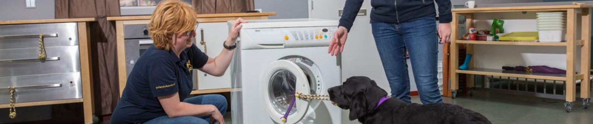 Marlouke en Tessa staan bij een wasmachine die door  een hond via een touwtje wordt opengetrokken