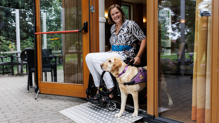 Mayke in een rolstoel samen met haar hulphond Roos in een deuropening