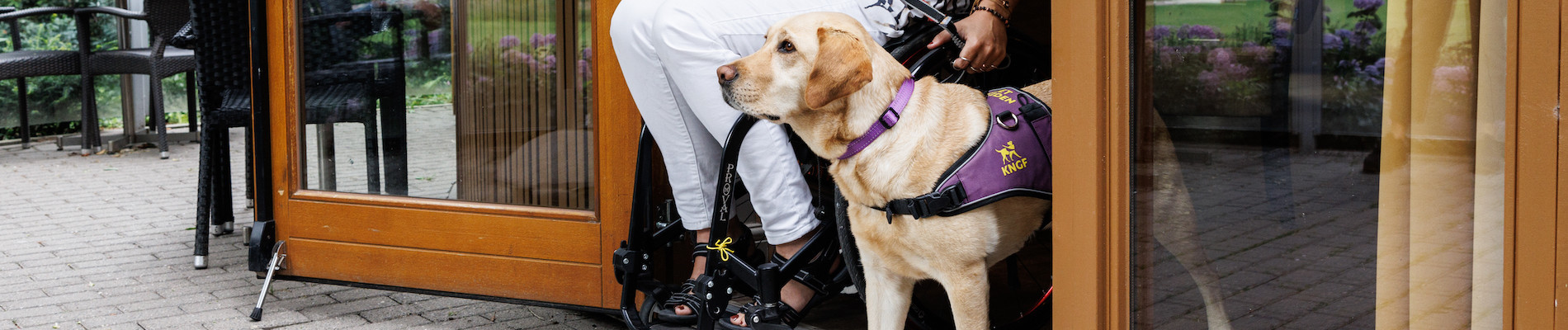 Mayke in een rolstoel samen met haar hulphond Roos in een deuropening