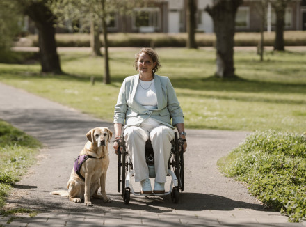 Vrouw in een rolstoel met een blonde hond in KNGF-tuig naast zich buiten in een park