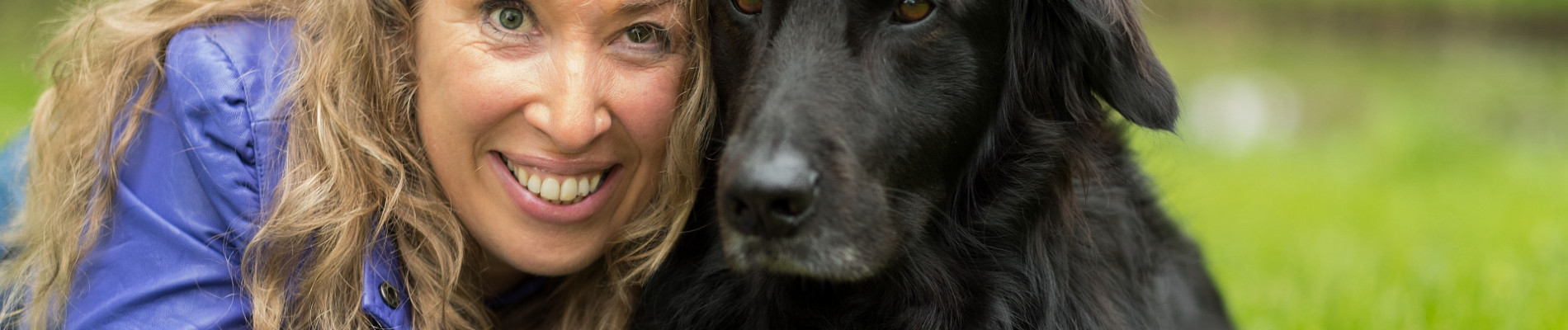 Petra en haar zwarte blindengeleidehond Wiggles liggen dicht naast elkaar in het gras en kijken de camera in.
