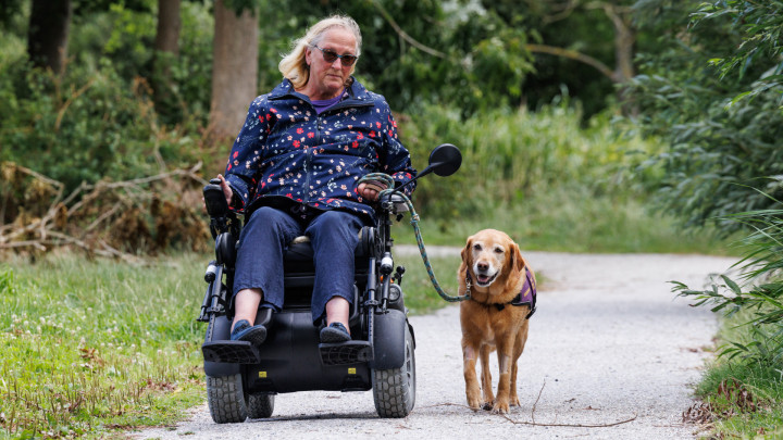 Mieke in de rolstoel buiten in het park, naast haar loopt ADL-assistentiehond Zonta