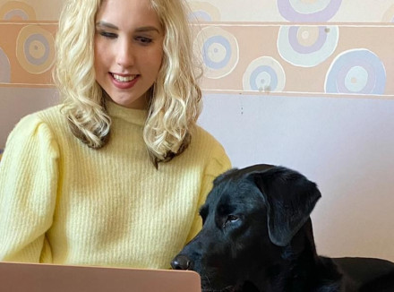 Sylvana zit op bed met een laptop op schoot en haar geleidehond ligt naast haar en kijkt mee naar het scherm