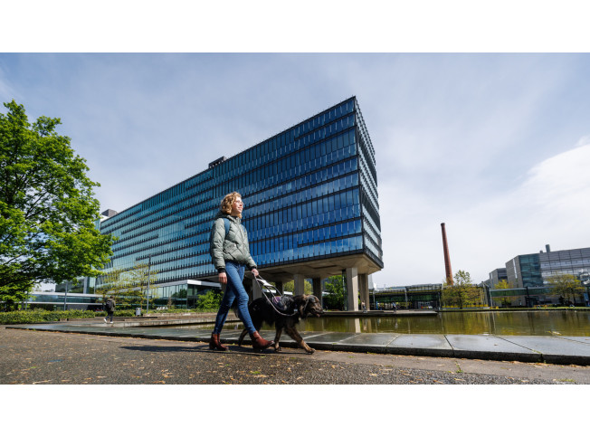 Femke loopt buiten met haar hond in tuig buiten voor een groot gebouw langs