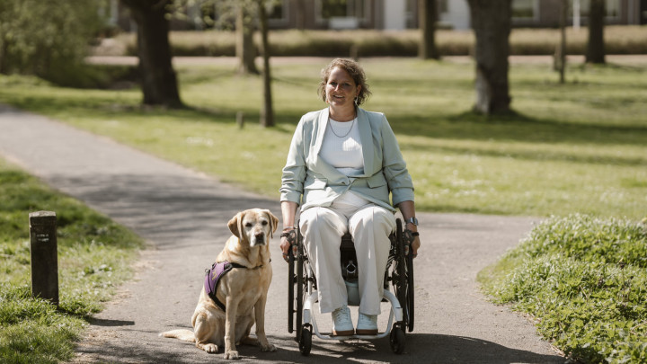 Vrouw in een rolstoel met een blonde hond in KNGF-tuig naast zich buiten in een park