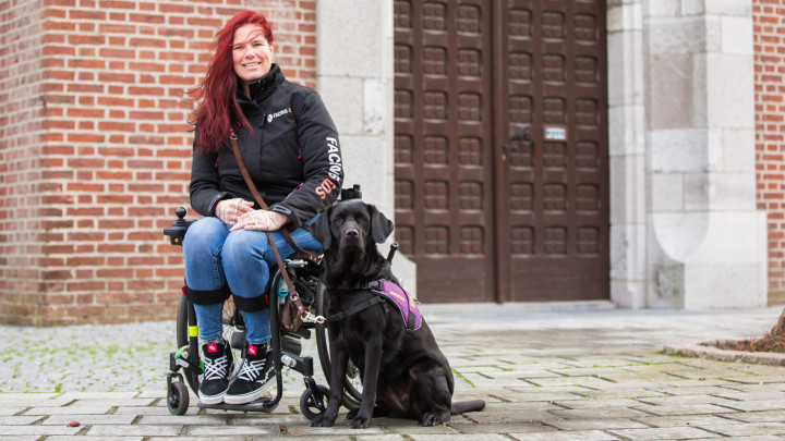 Mariska zit in een rolstoel buiten voor een gebouw met een grote deur. Haar zwarte assistentiehond Aura zit naast haar