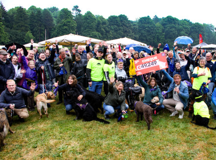 Groepsfoto van mensen en honden. In het midden directeur Monique Dolfing en Jamie Trenité die een grote cheque omhoog houden.