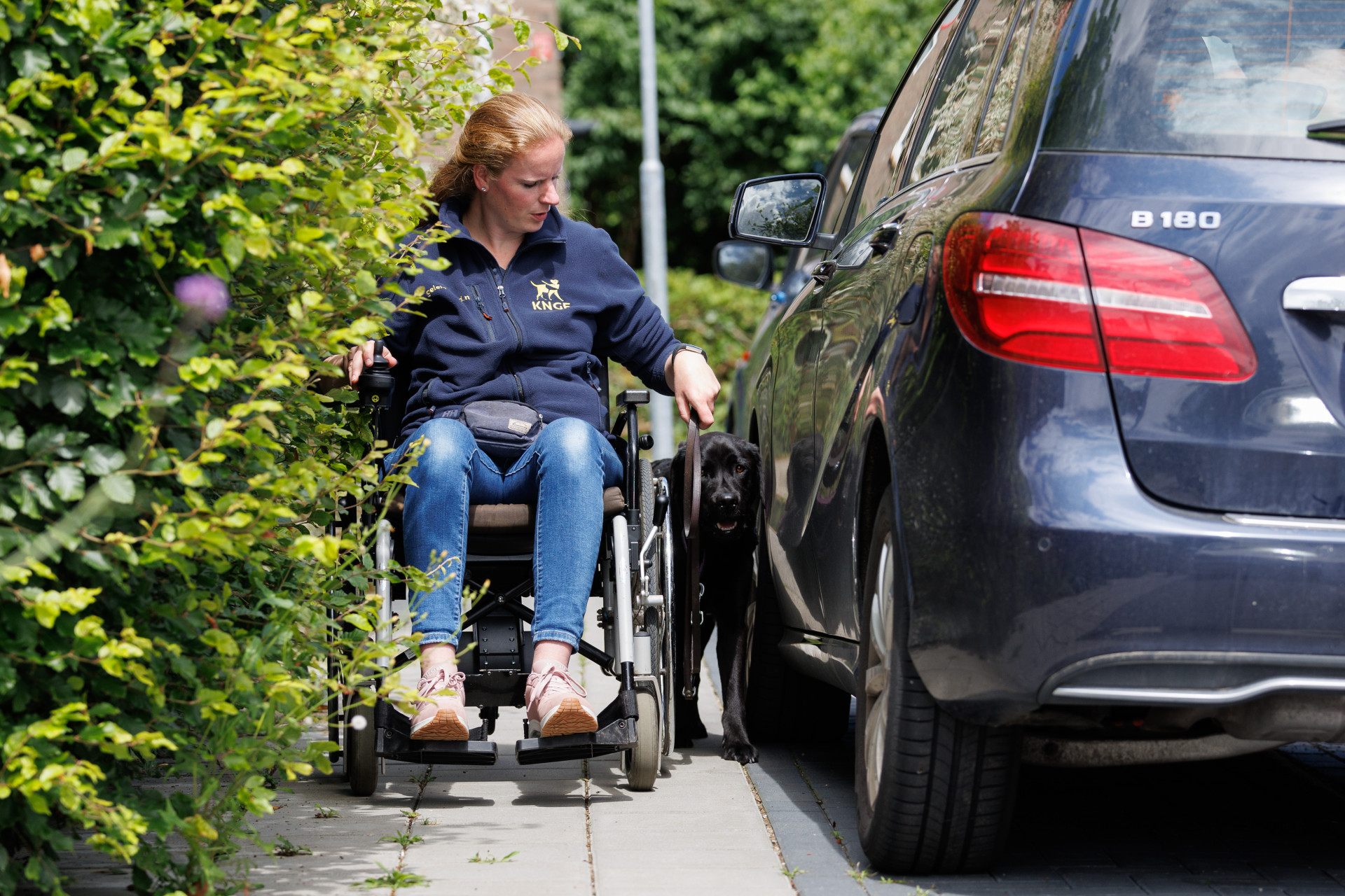 Tessa rijdt op de krappe stoep in een rolstoel tussen auto en heg.