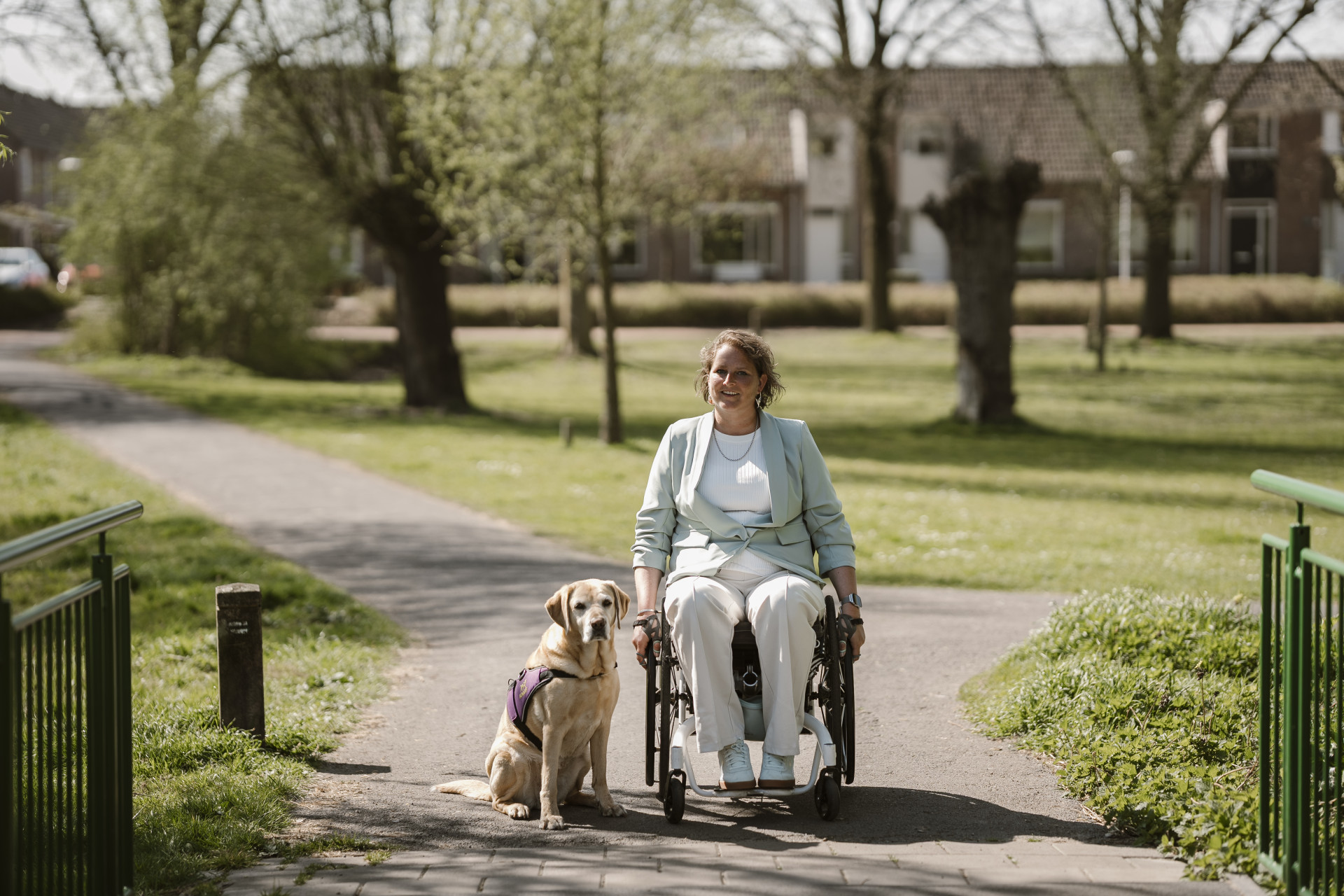 Vrouw in een rolstoel met een blonde hond in KNGF-tuig naast zich buiten in een park