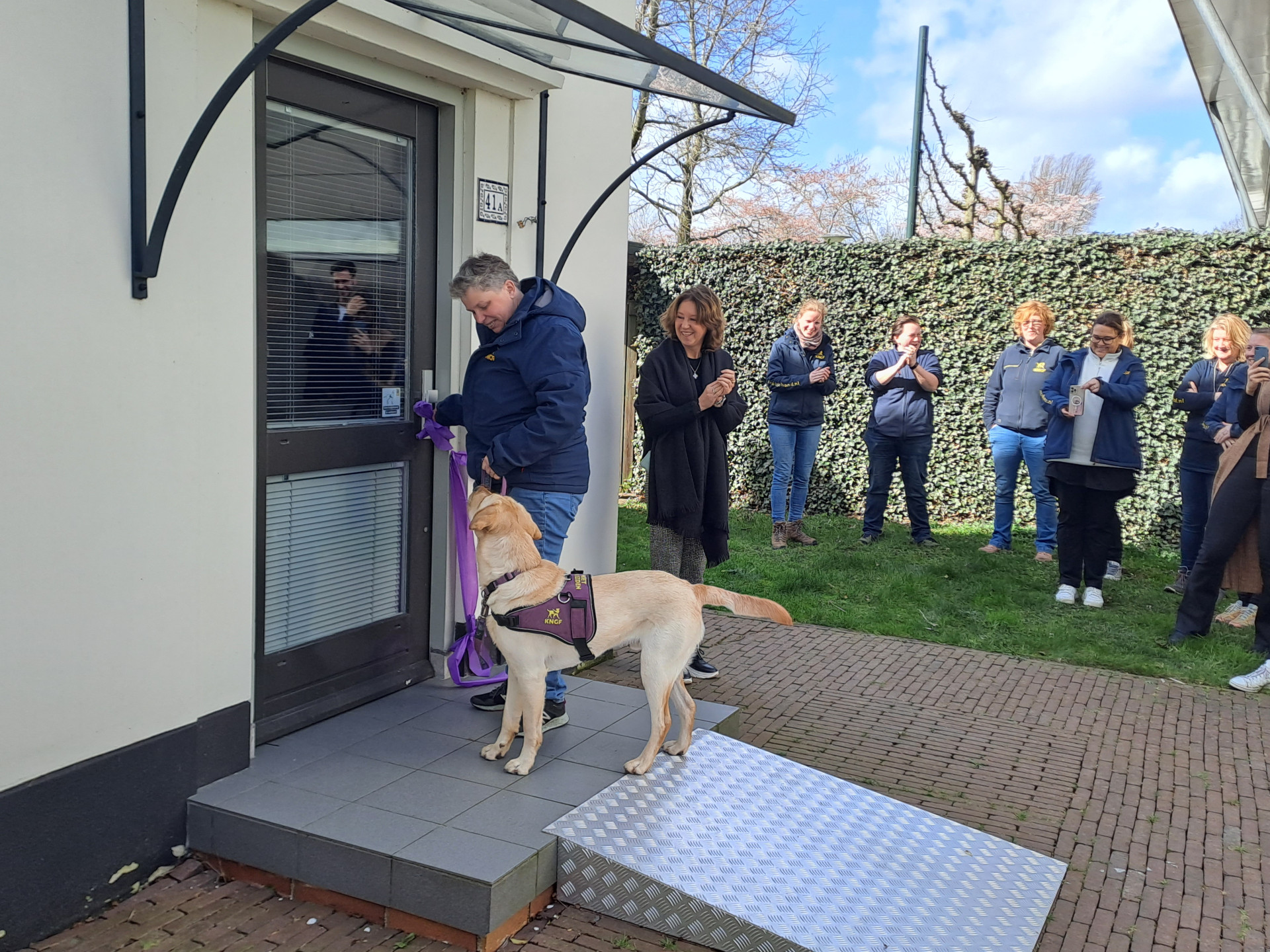 blonde hond staat voor de voordeur van het trainingshuis. Het paarse lint hangt nog aan de deur. Eromheen staat collega's