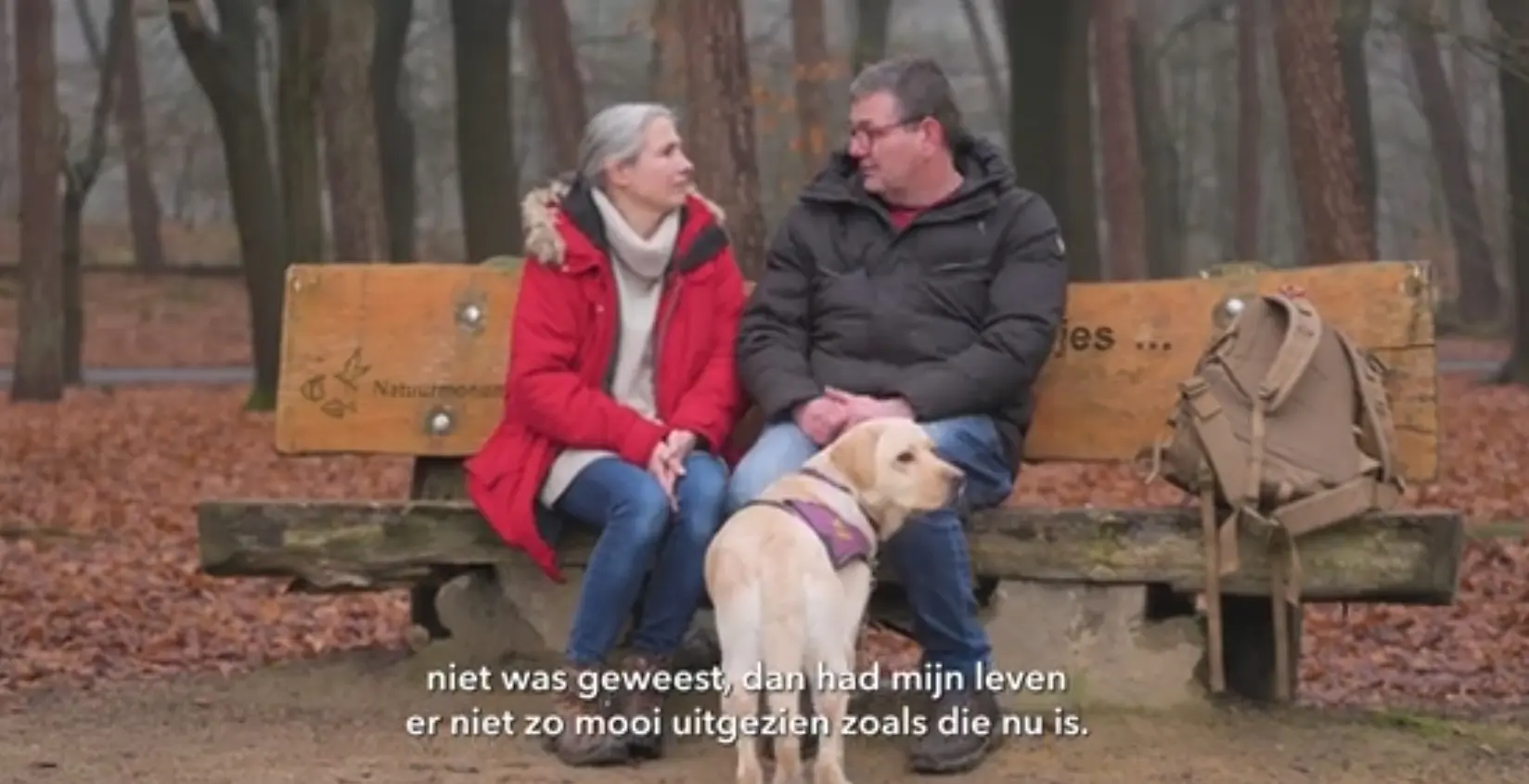 Cristel en Edwin zitten naast elkaar op een bank in het bos te praten. Een blonde hond staat voor Edwin.