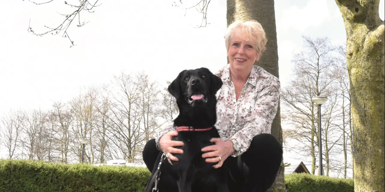 Vrijwilliger Ria zit op het gras met een zwarte labrador naast haar