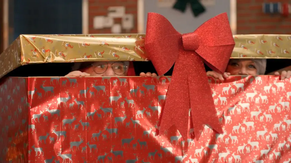 2 collega's zitten in een enorme cadeauverpakking en spieken onder de deksel naar buiten.