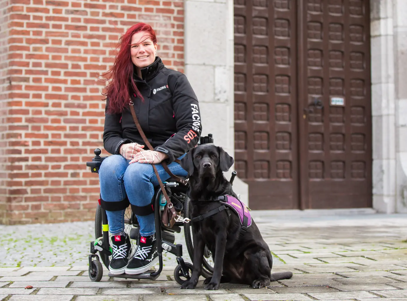 Mariska zit in een rolstoel buiten voor een gebouw met een grote deur. Haar zwarte assistentiehond Aura zit naast haar