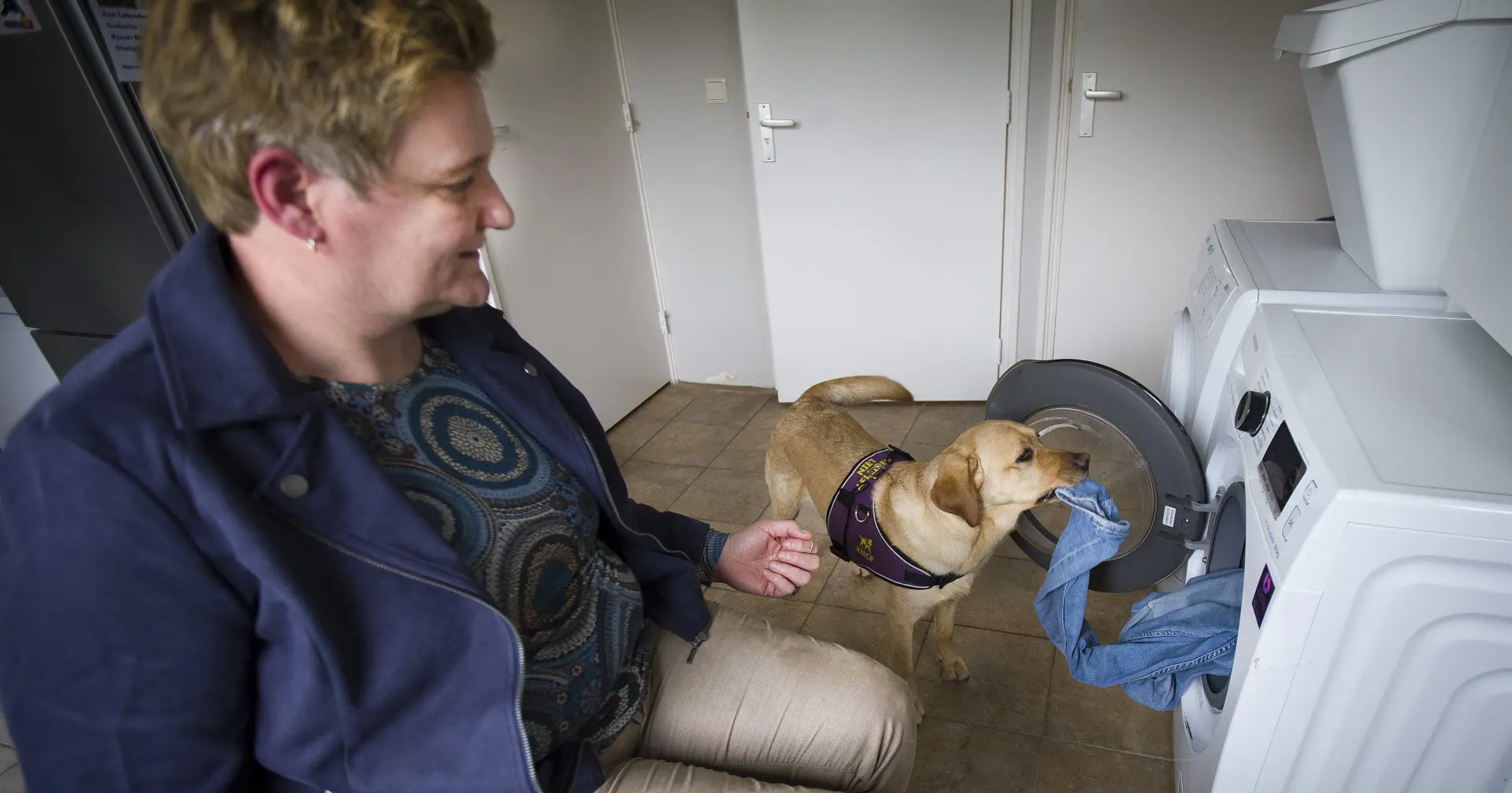 Assistentiehond Danja haalt de was uit de wasmachine terwijl Cindy toekijkt
