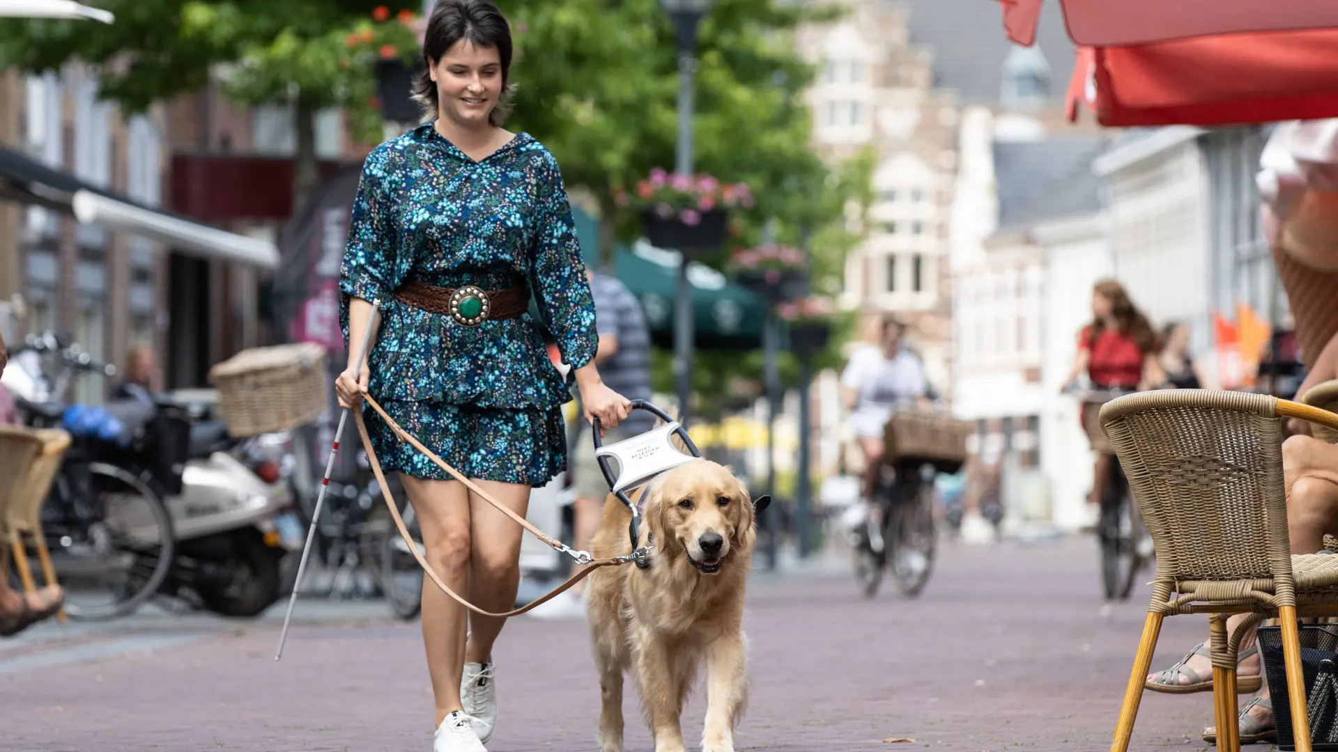 Lisa, in een vrolijke jurk, loopt samen met haar blindengeleidehond Boyka over straat langs een terras