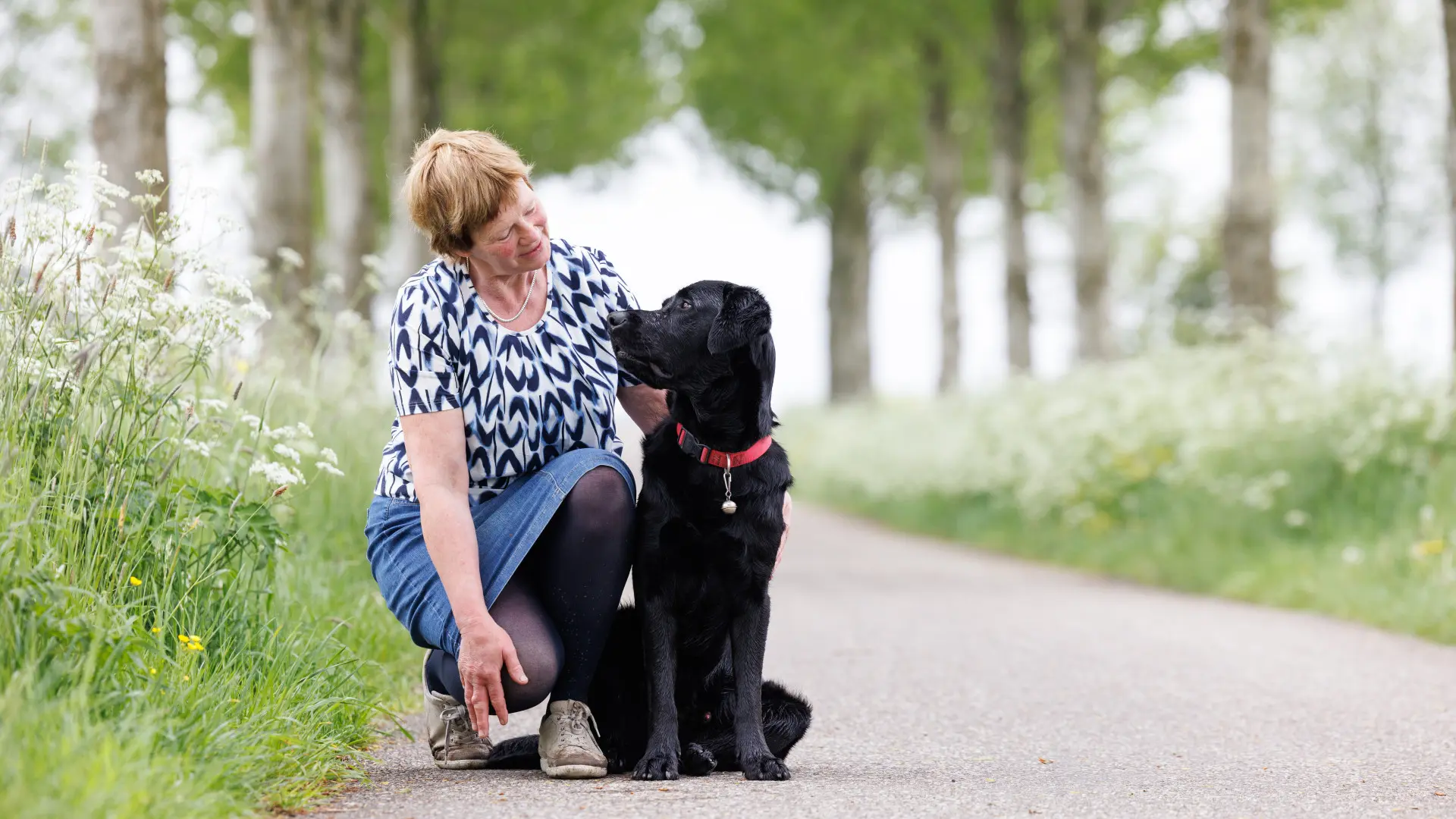 Clary hurkt op de straat naast haar blindengeleidehond Flipse, een zwarte labrador kruising.