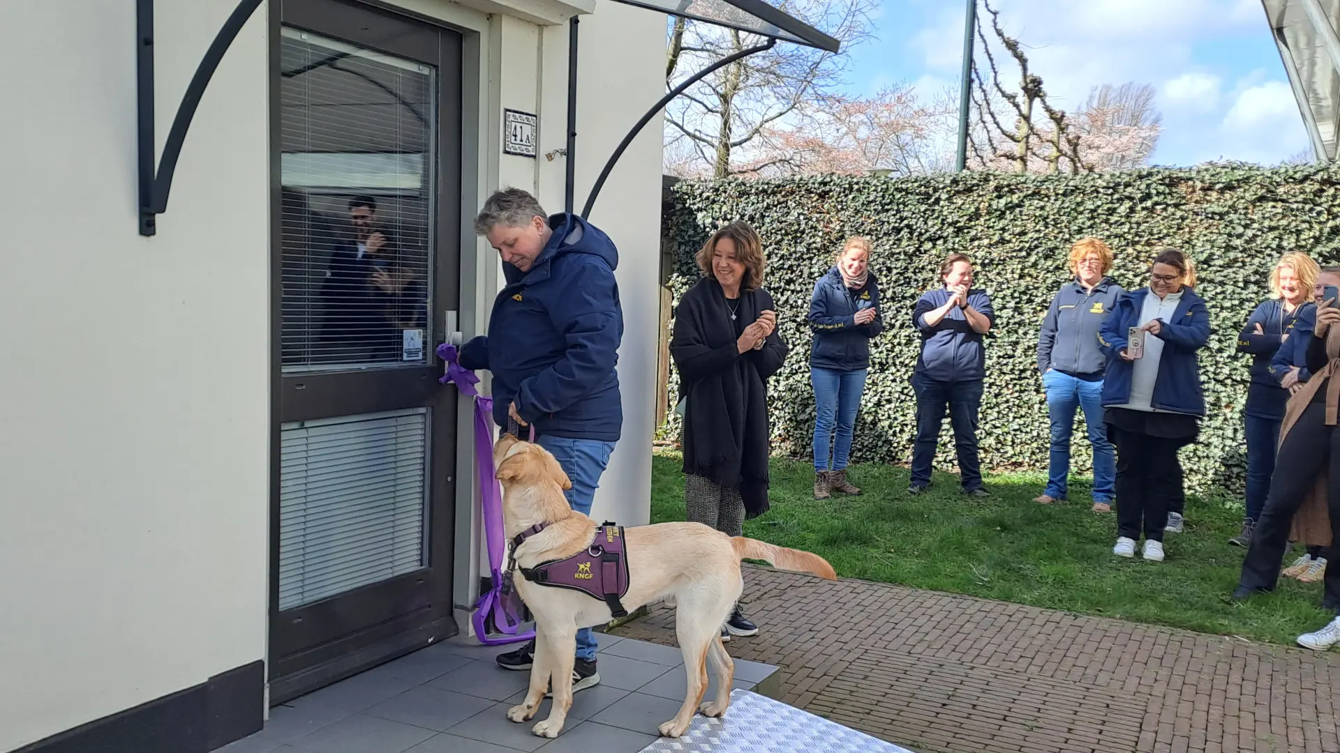 Onder toeziend oog van genodigden en trainster Annemieke opent een assistentiehond het trainingshuis