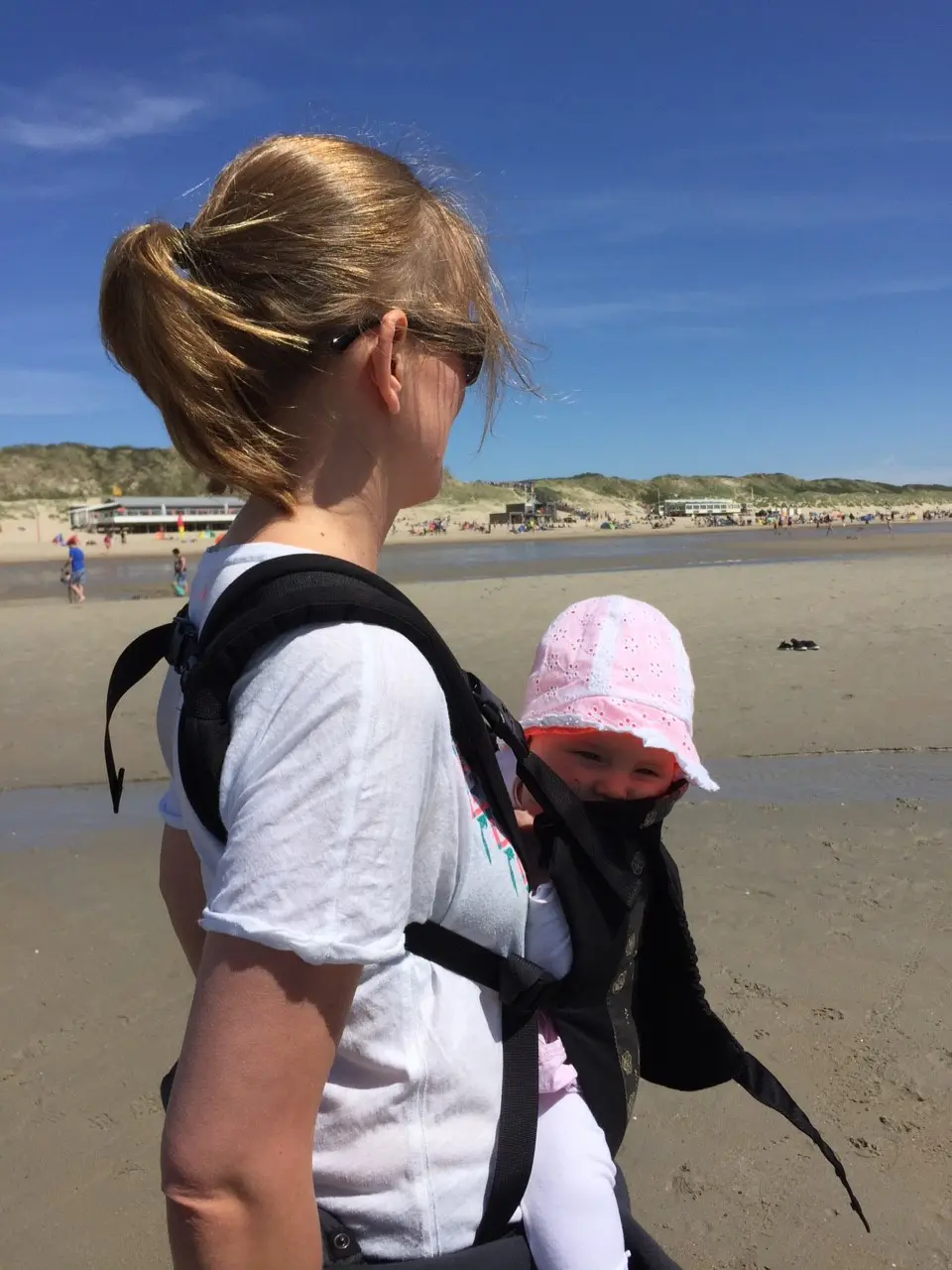 Anneloes kijkt uit op het strand met haar baby in een draagzak op haar buik