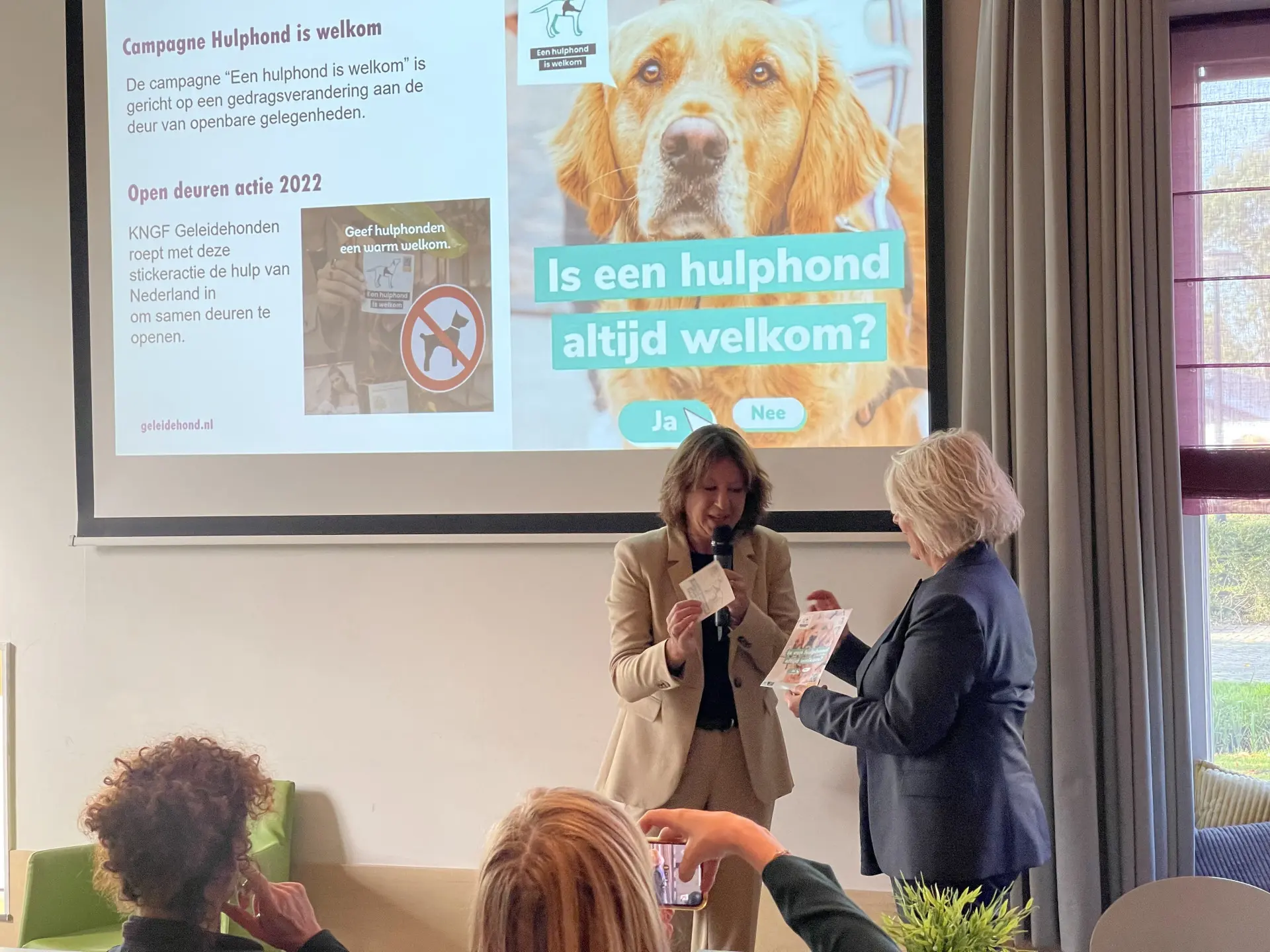 Secretaris-generaal bij OCW Marjan Hammersma krijgt van KNGF directeur Monique Dolfing de campagnesticker overhandigd met ‘een hulphond is welkom’.