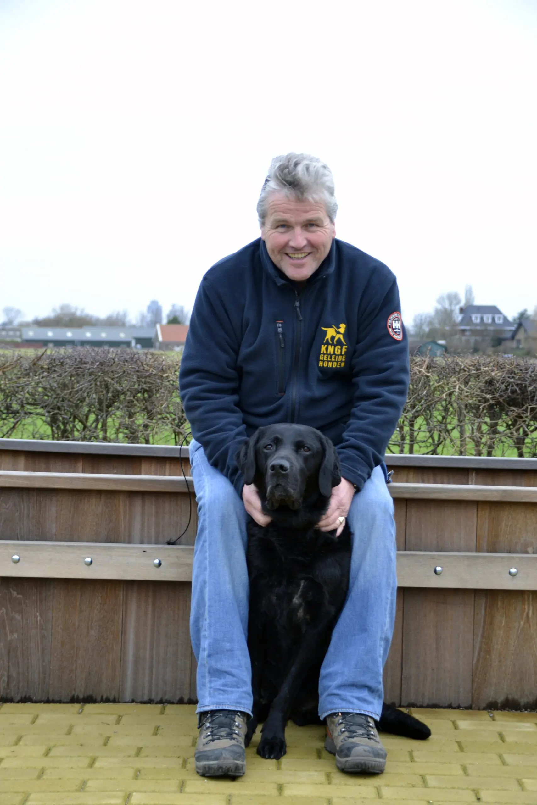 Trainer Kees zit op een houten verhoging met voor zich een zwarte labrador