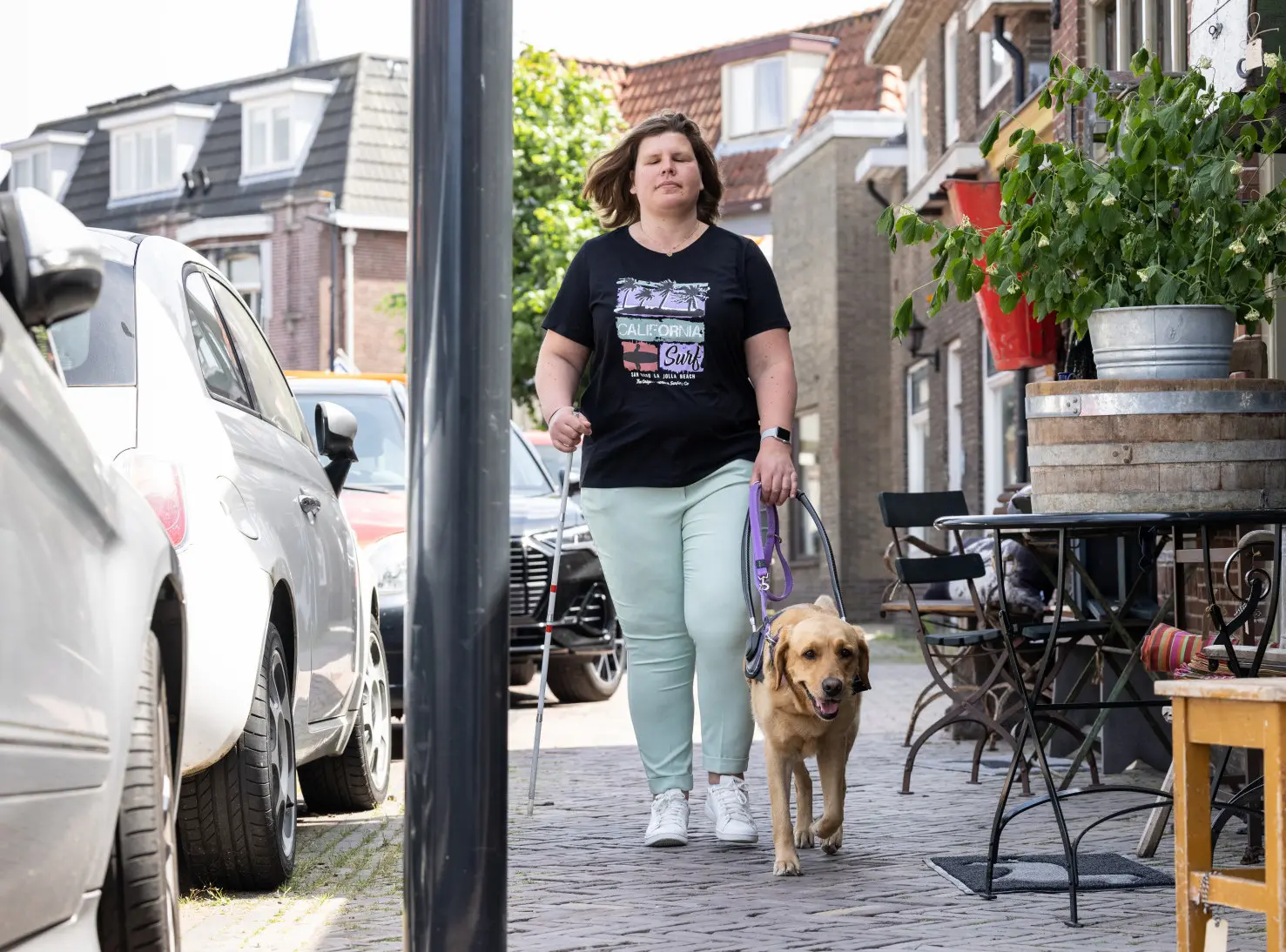 Melanie en haar blindengeleidehond Ajonne lopen over straat met winkels en geparkeerde autos naast hun