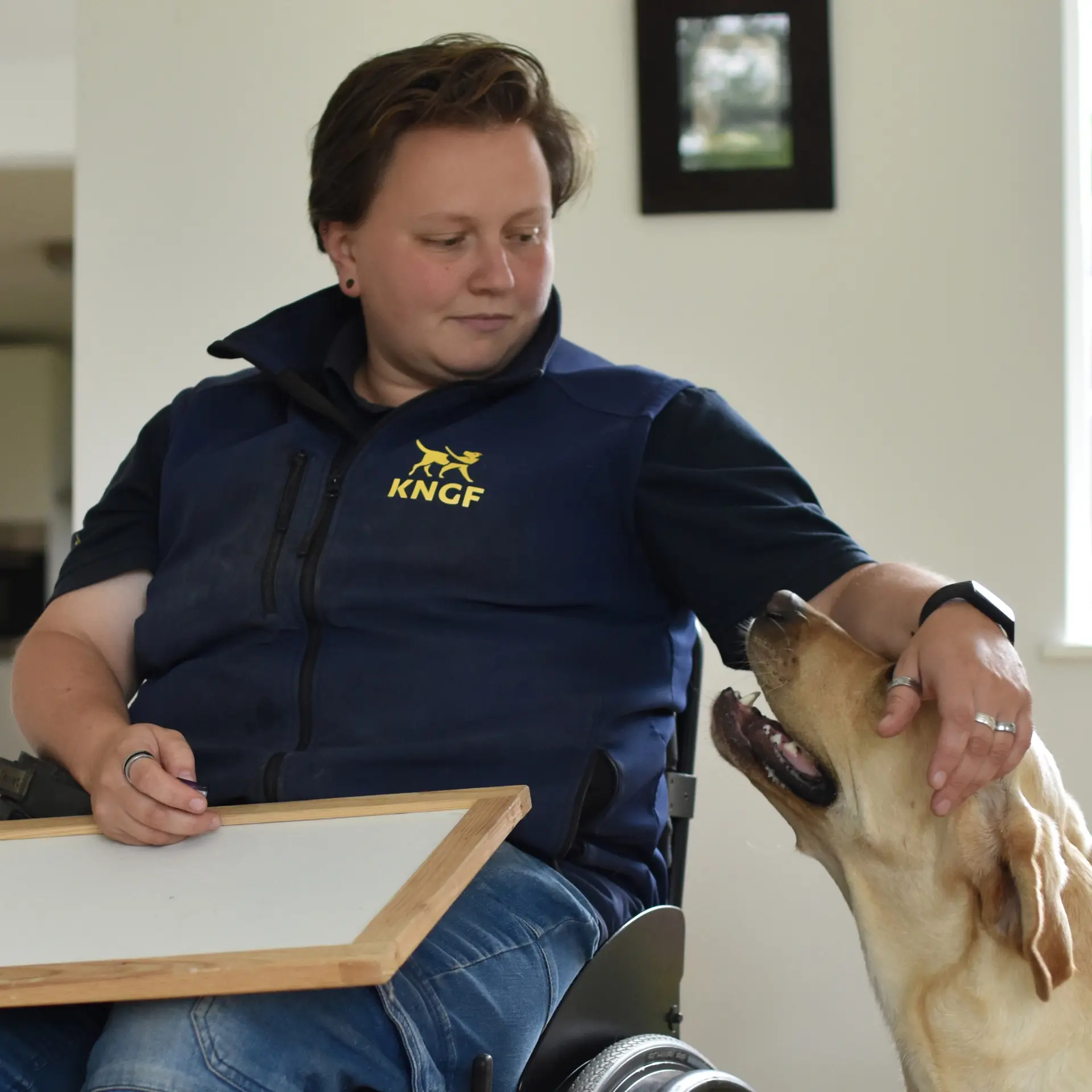 Trainster Priscilla geeft een assistentiehond een aai over de kop terwijl ze in een rolstoel achter een tafel zit