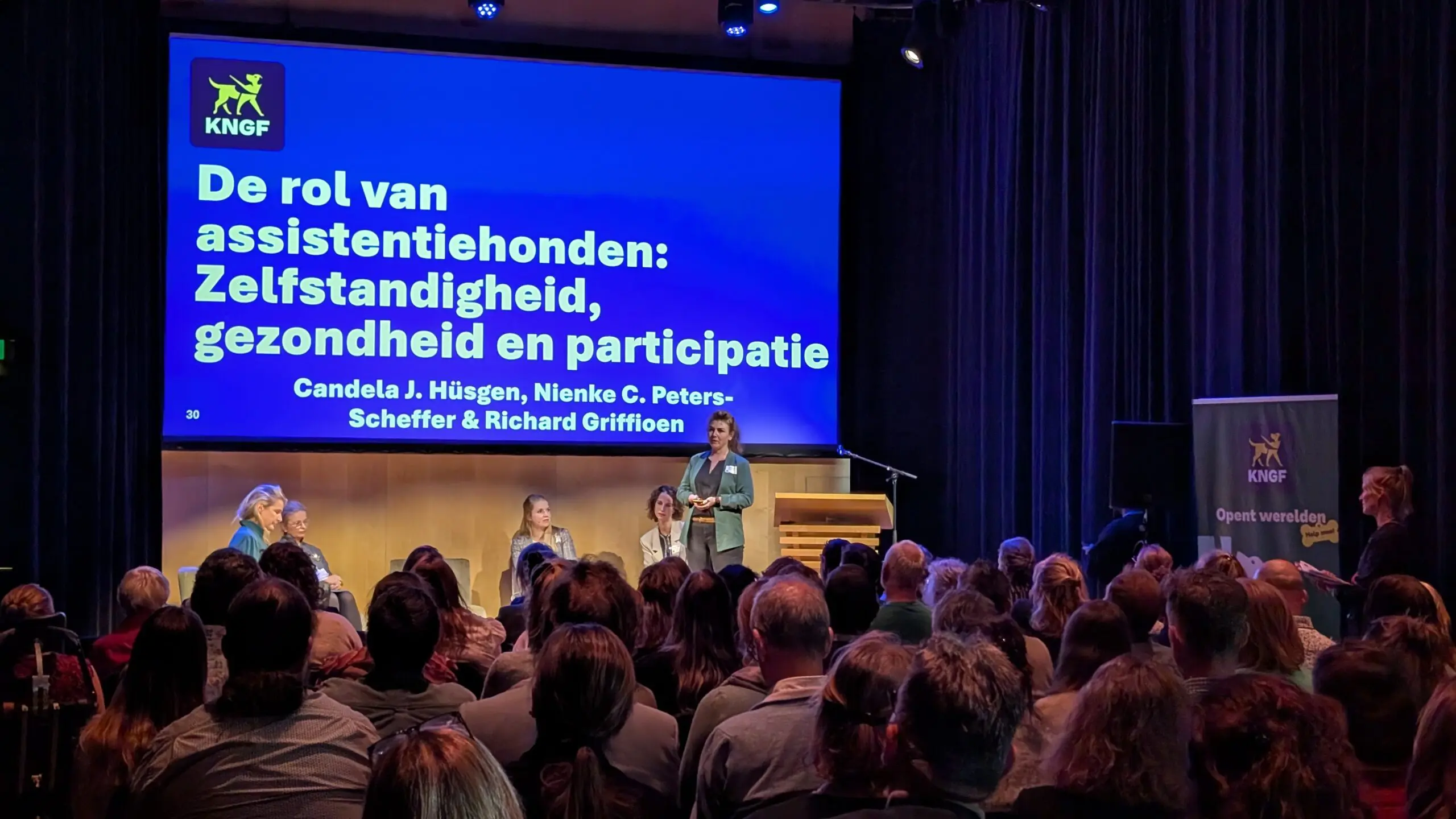 een vrouw geeft een presentatie voor een zaal met toehoorders. Achter haar een presentatiescherm met tekst.