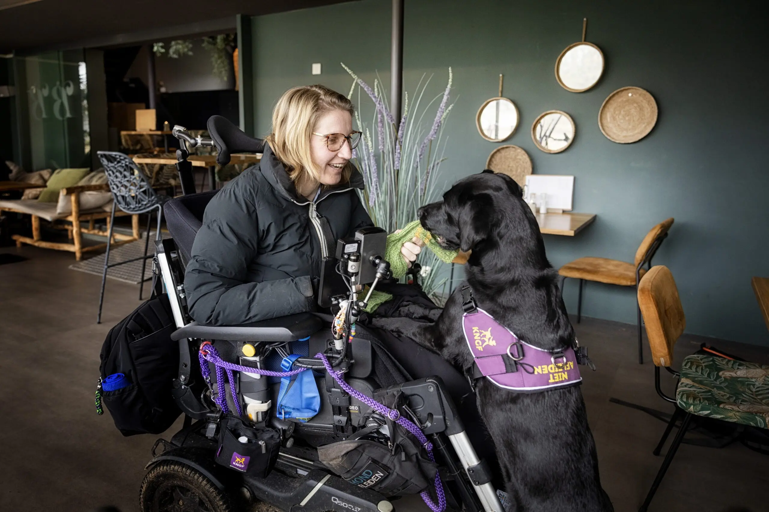 Vrouw in rolstoel in huiselijke omgeving, haar zwarte assistentiehond staat met haar poten op haar schoot