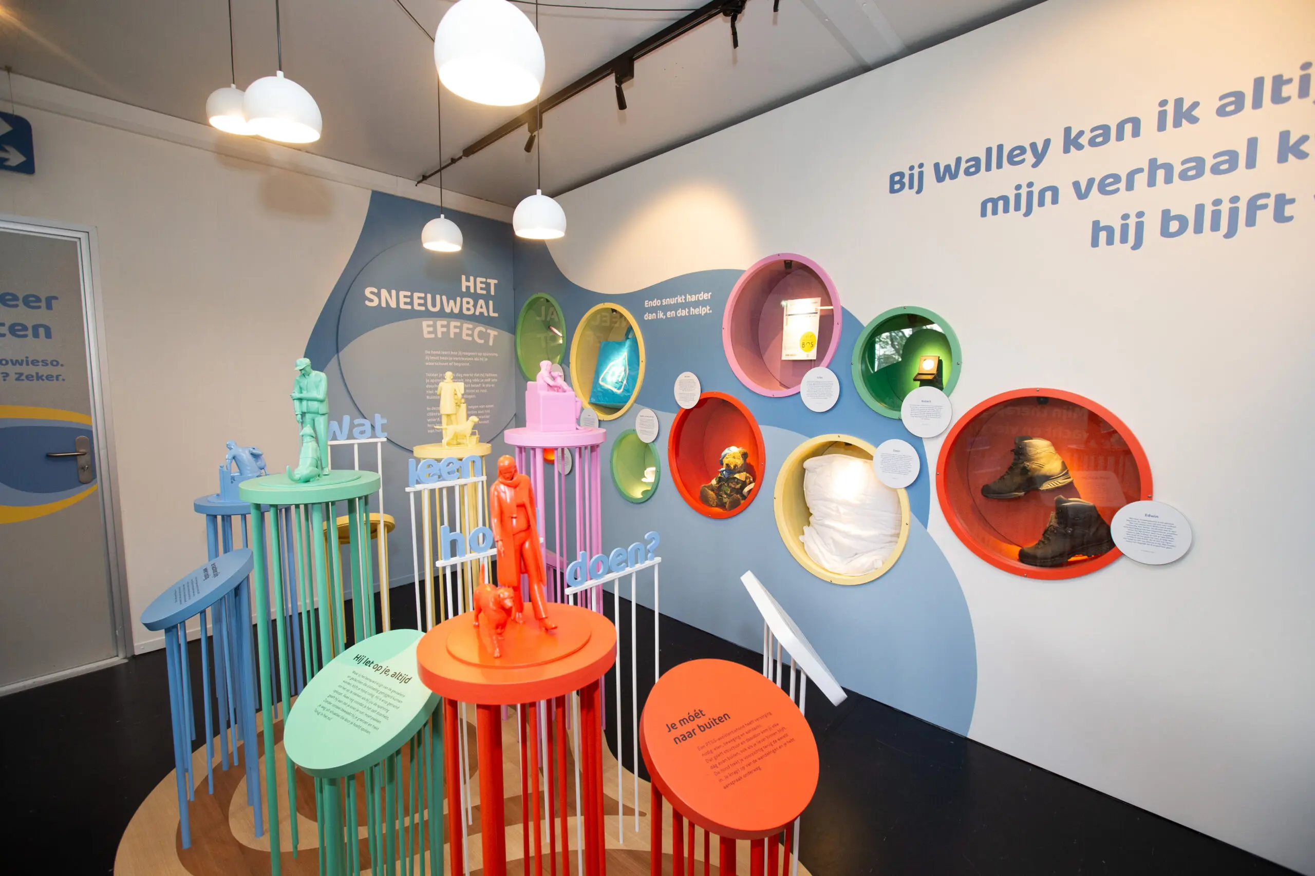 Kleurig beeld van pilaren met daarop een tekst of beeld, op de achtergrond teksten op de muur en uitstallingen van verschillende objecten zoals een knuffel en schoenen.