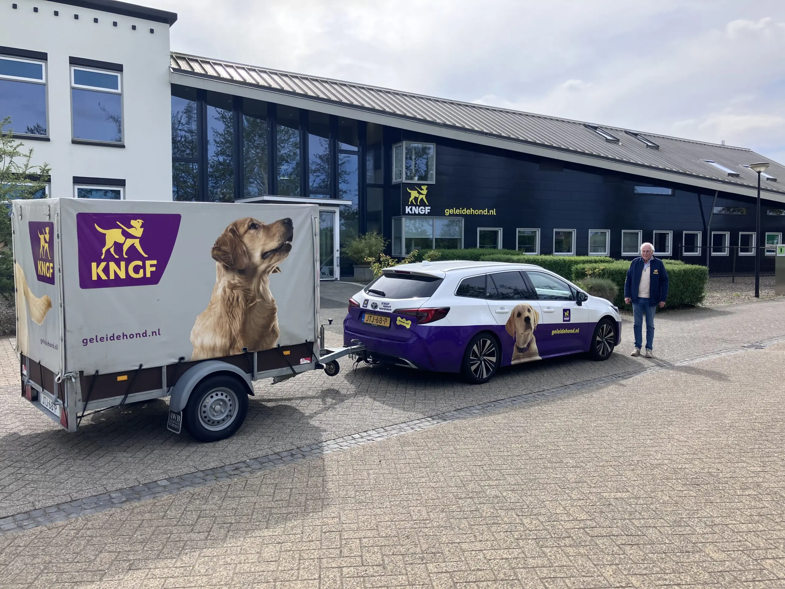 man staat voor het KNGF-hoofdgebouw met een auto in KNGF-kleuren en erachter aan aanhanger.