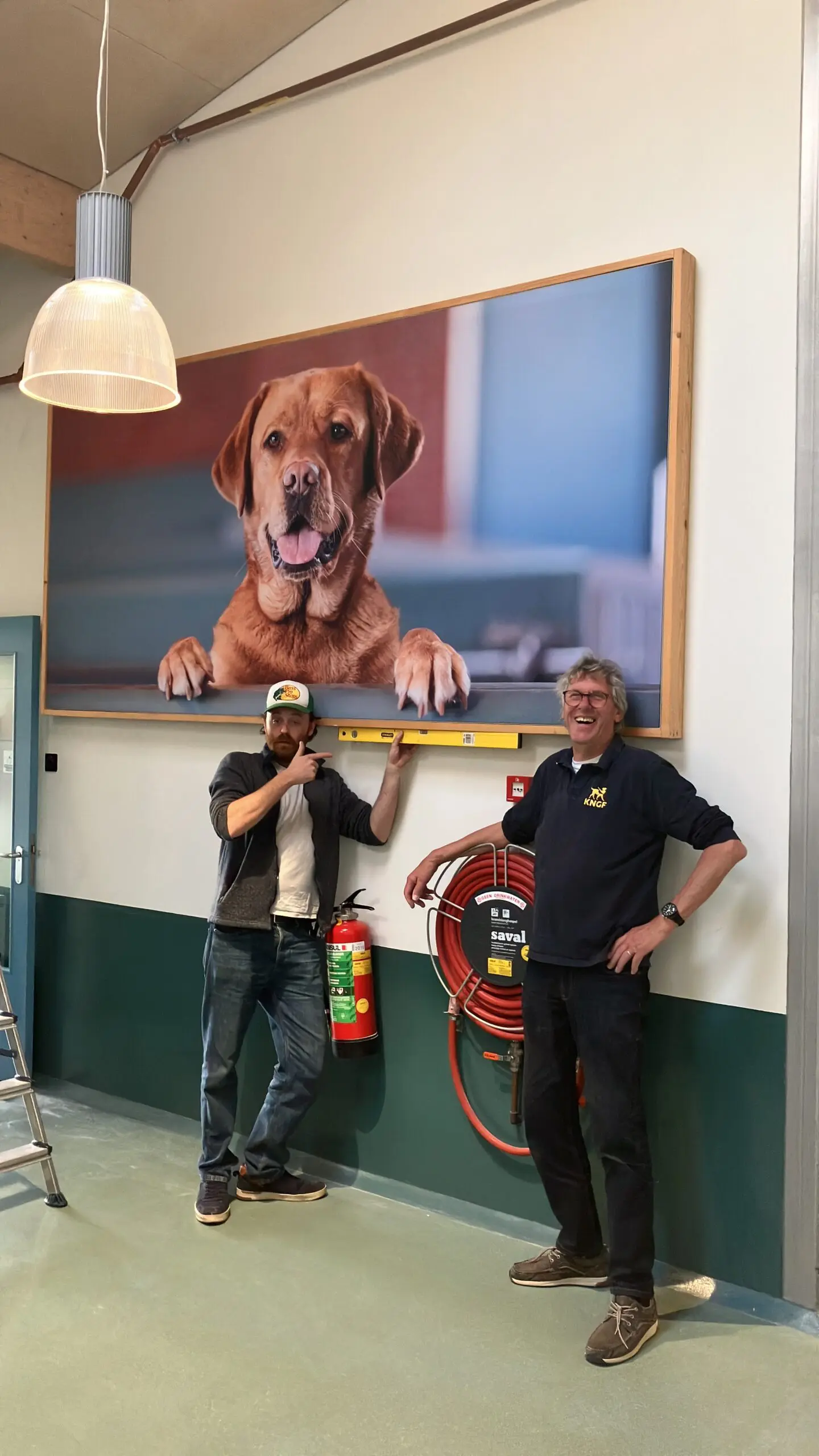 2 mannen staan onder een grote portretfoto van een hond en houden er een waterpas onder.