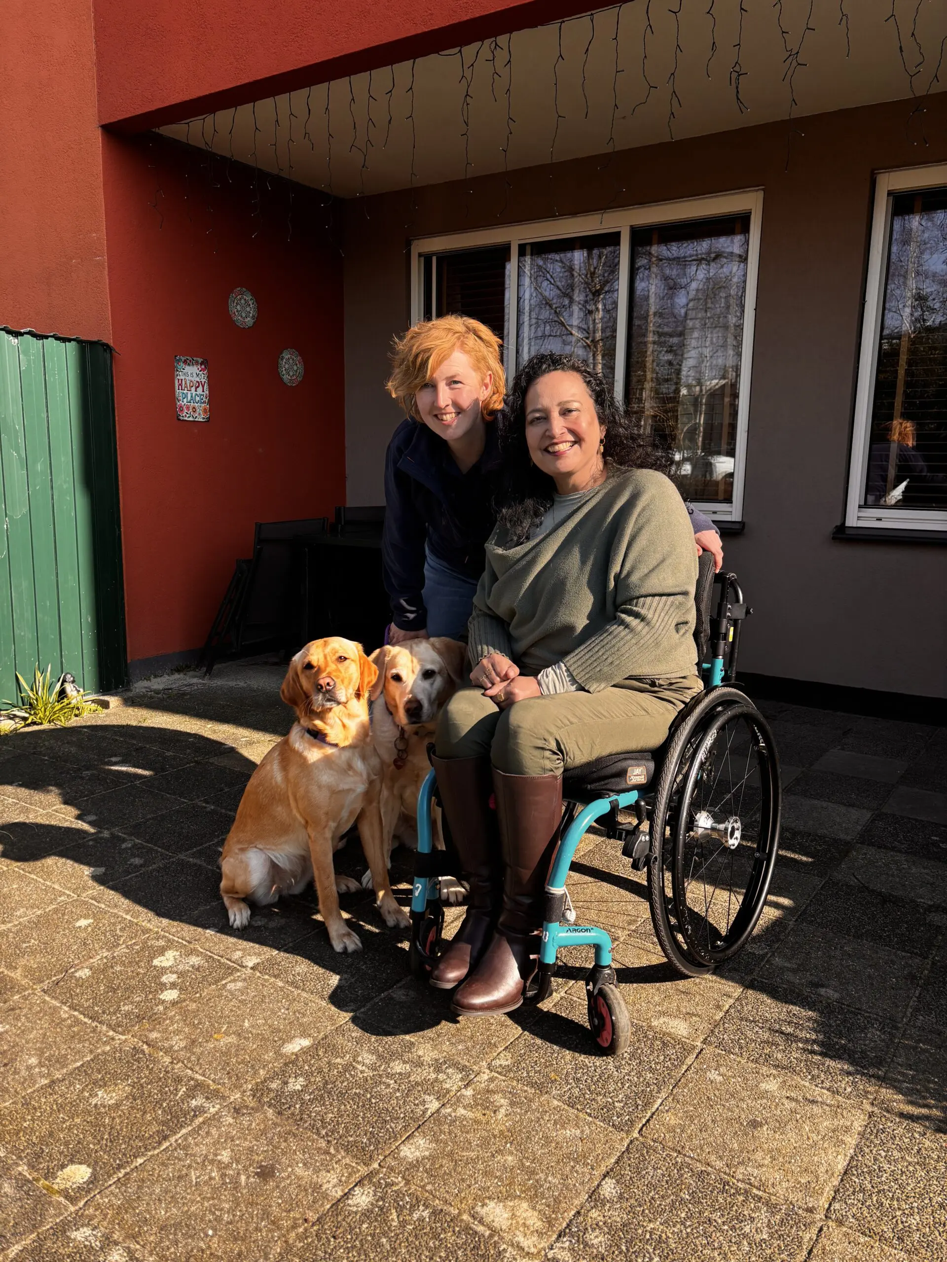 Claudia samen met instructeur Marlouke en naast haar rolstoel 2 blonde honden die naast elkaar zitten.