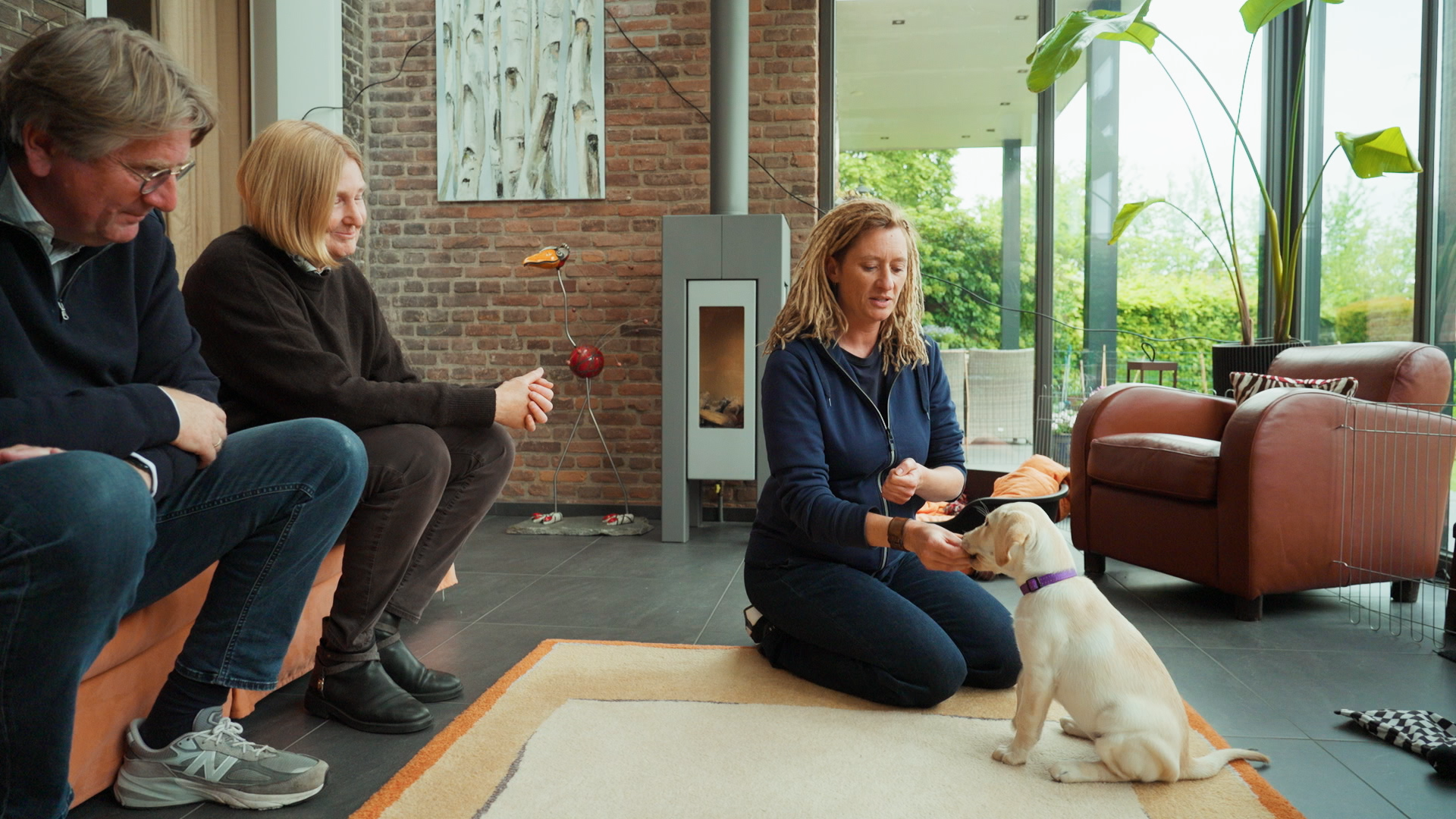 puppyinstructeur zit in woonkamer op de grond en geeft een kleine zittende blonde pup een voertje. Naast haar zitten een man en een vrouw op de bank die meekijken.