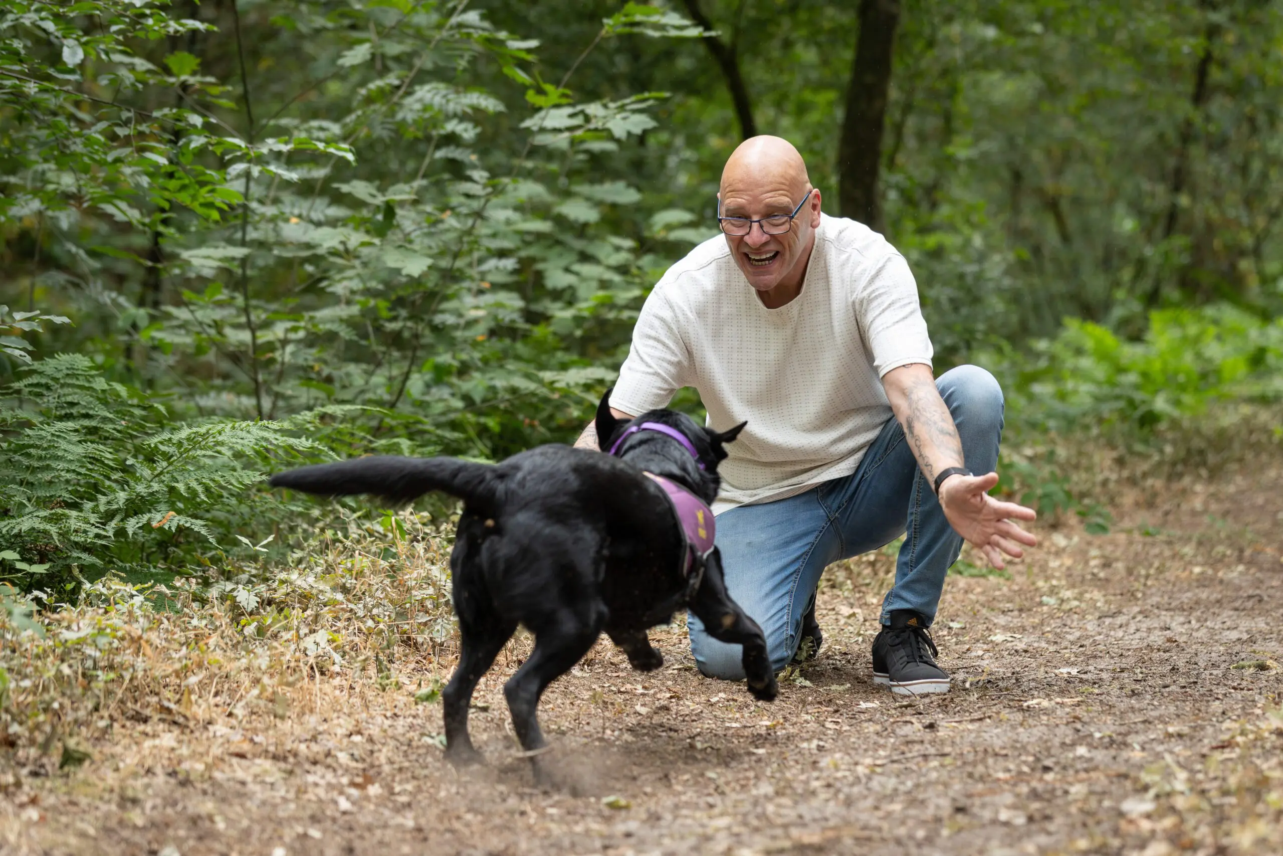 Ron zit op zijn knieën in het bos en zijn zwarte labrador Becky komt op hem af rennen.