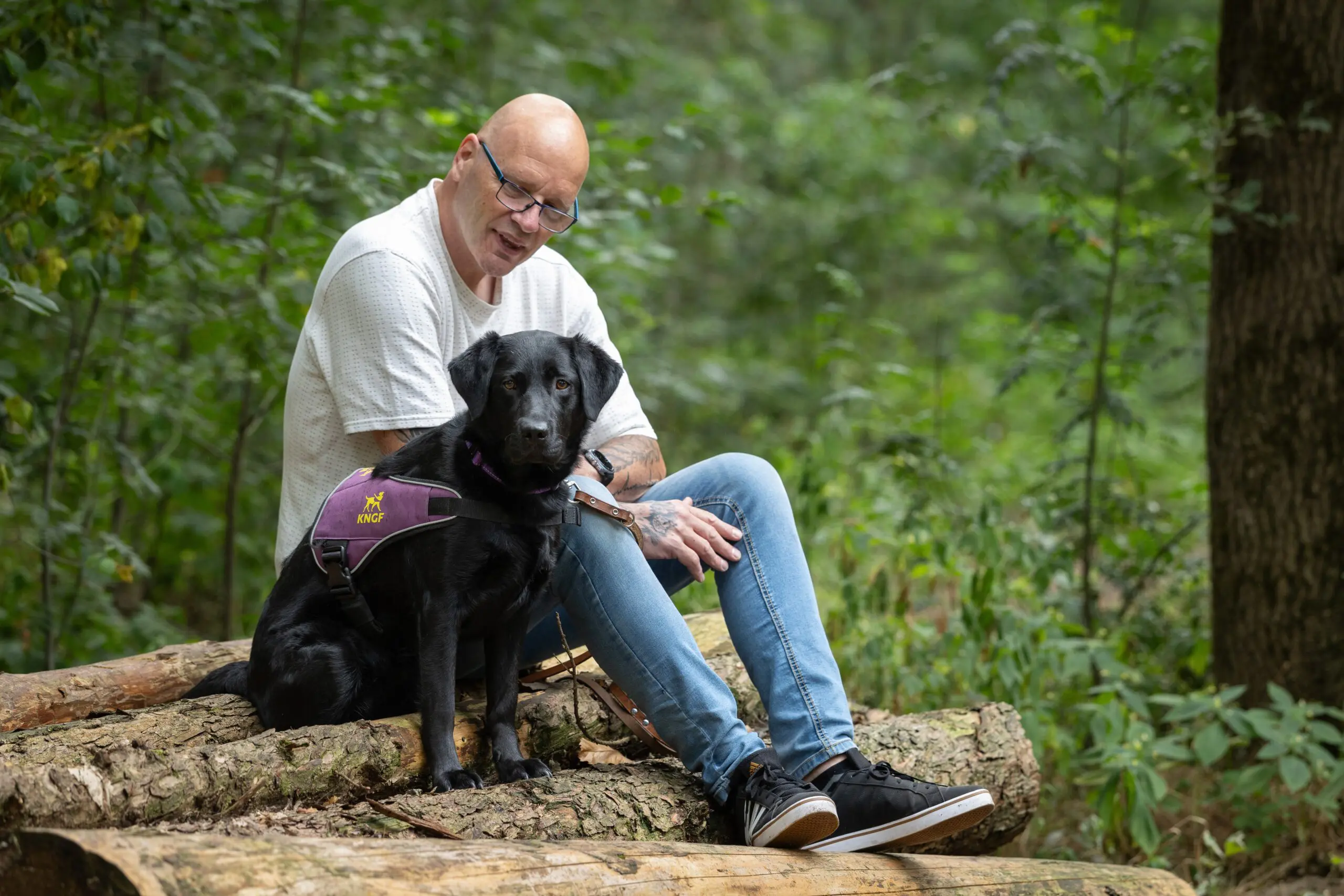 Ron zit op een stapel boomstammen met zijn zwarte labrador Becky naast zich