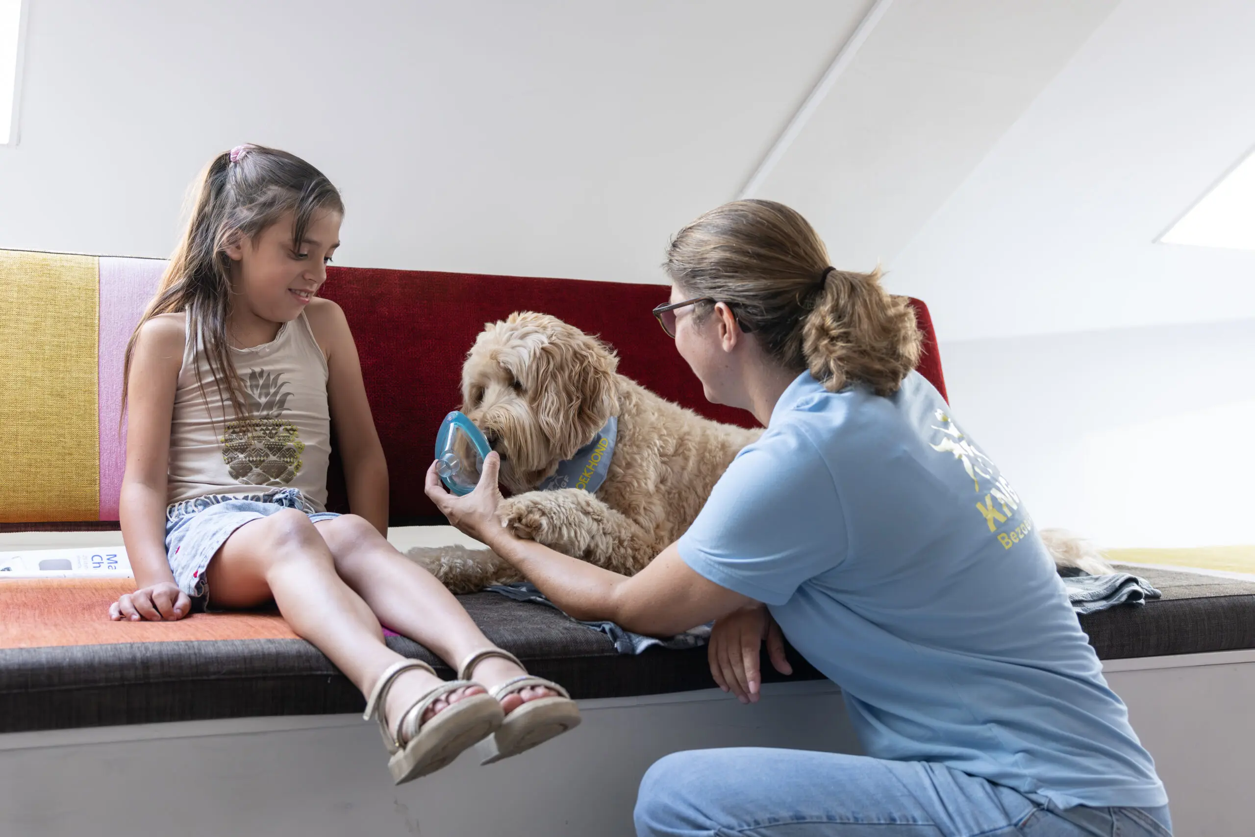 Meisje Rosalyn en bezoekhond Flynn zitten op een bakje. Begeleider Laura laat zien hoe Flynn aan een narcosekapje snuffelt