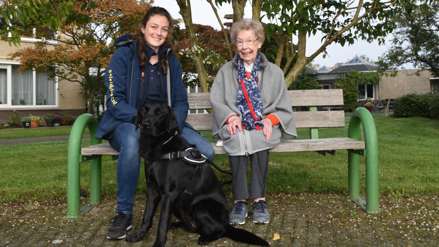 Donateur Frida zit op bankje naast trainer KNGF en hond in opleiding