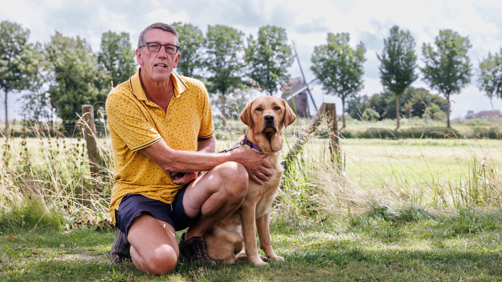 Jan Combee staat samen met jonge hulphond op de foto met op de achtergrond een graslandschap met een molen in de verte